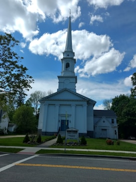 A white church with a tall steeple is set against a vibrant blue sky filled with scattered clouds. The building features classical architectural elements, including columns and a triangular pediment. Surrounding the church are lush green trees and a well-maintained lawn with some flowers. A sign in front of the church provides information about upcoming events.
