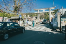 A traditional Japanese street featuring a large torii gate. A taxi is parked on the left side, and several signs written in Japanese are visible. The sky is clear and blue, with a few trees scattered around the scene.