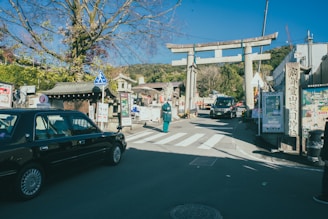 A traditional Japanese street featuring a large torii gate. A taxi is parked on the left side, and several signs written in Japanese are visible. The sky is clear and blue, with a few trees scattered around the scene.