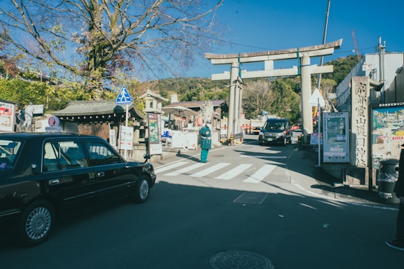 A traditional Japanese street featuring a large torii gate. A taxi is parked on the left side, and several signs written in Japanese are visible. The sky is clear and blue, with a few trees scattered around the scene.