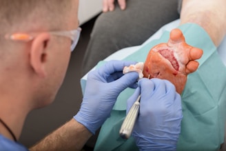 A medical professional wearing gloves performs a surgical procedure on a foot. The foot has visible incisions and exposed tissue, indicating a complex surgical intervention. The medical professional is using precise tools to work on the area, suggesting a medical or surgical setting.