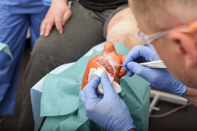 A medical professional wearing blue gloves is performing a procedure on a wounded hand. The hand appears to have a significant injury, and the professional is using tools to attend to it. The patient is seated, and surgical drapes cover parts of the hand and surrounding area.