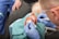 A medical professional wearing blue gloves is performing a procedure on a wounded hand. The hand appears to have a significant injury, and the professional is using tools to attend to it. The patient is seated, and surgical drapes cover parts of the hand and surrounding area.
