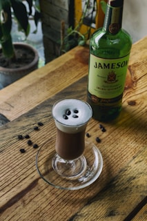 A wooden table adorned with a glass containing a frothy coffee beverage, topped with coffee beans. A bottle of Jameson Irish Whiskey is positioned next to the glass. Coffee beans are scattered on the table, enhancing the rustic aesthetic. A potted plant and a window in the background contribute to a cozy atmosphere.
