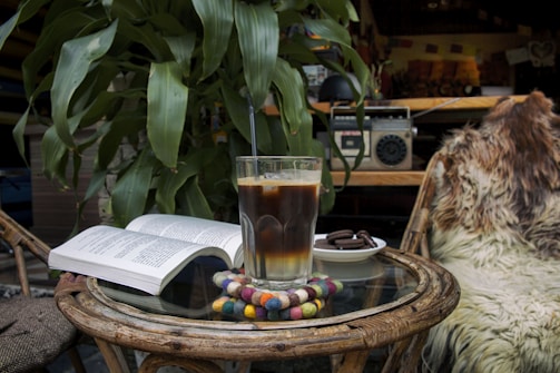 A cozy setting with a glass of iced coffee placed on a circular wicker table. A colorful coaster is underneath the glass, and there is an open book nearby. A small plate with cookies is next to the drink. In the background, a large leafy plant adds greenery, and an old-fashioned radio is visible. A furry blanket is draped over a chair, enhancing the warm atmosphere.