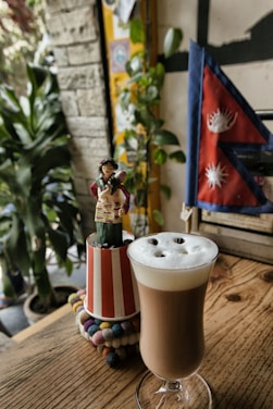 A glass of frothy coffee sits on a wooden table, surrounded by colorful decor including a small doll figurine and a vibrant flag with a sun and moon design. The setting appears cozy and inviting with plants adding greenery in the background.