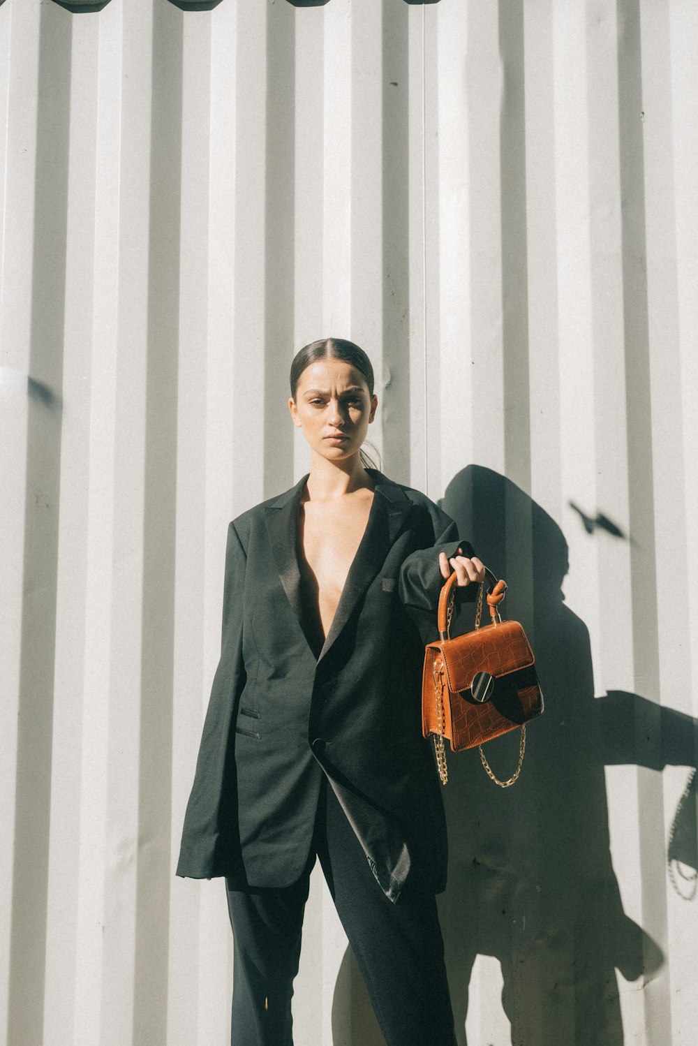 woman wearing black blazer holding brown handbag leaning on white wall