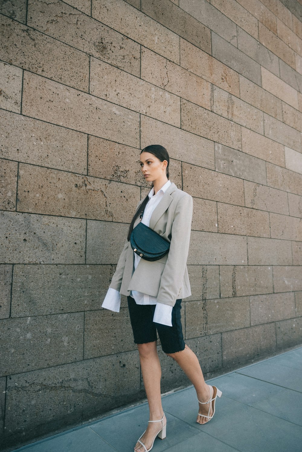 woman in beige blazer, black shorts, and pair of chunky heeled sandals walking near wall