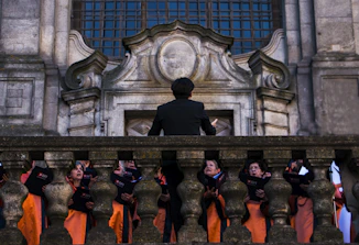 A group of choral singers dressed in orange robes is gathered on a grand stone staircase, led by a conductor standing with his back to the camera. The ornate stone architecture suggests a historical or religious setting. The singers hold black folders and appear to be deeply focused, possibly in the middle of a performance.