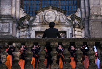 A group of choral singers dressed in orange robes is gathered on a grand stone staircase, led by a conductor standing with his back to the camera. The ornate stone architecture suggests a historical or religious setting. The singers hold black folders and appear to be deeply focused, possibly in the middle of a performance.