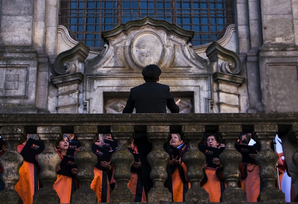 A group of choral singers dressed in orange robes is gathered on a grand stone staircase, led by a conductor standing with his back to the camera. The ornate stone architecture suggests a historical or religious setting. The singers hold black folders and appear to be deeply focused, possibly in the middle of a performance.