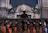 A group of choral singers dressed in orange robes is gathered on a grand stone staircase, led by a conductor standing with his back to the camera. The ornate stone architecture suggests a historical or religious setting. The singers hold black folders and appear to be deeply focused, possibly in the middle of a performance.