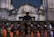 A group of choral singers dressed in orange robes is gathered on a grand stone staircase, led by a conductor standing with his back to the camera. The ornate stone architecture suggests a historical or religious setting. The singers hold black folders and appear to be deeply focused, possibly in the middle of a performance.