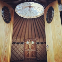 The interior of a circular yurt is visible, featuring wooden doors with round windows. The ceiling is designed with a radial wooden pattern, leading up to a round skylight at the top. The walls are lined with fabric and a lattice wooden framework.