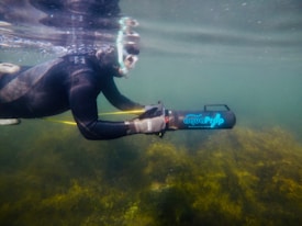 A person wearing a black wetsuit and snorkeling mask holds a device labeled 'aquaProp' underwater. The water is murky with visible marine vegetation on the bottom.