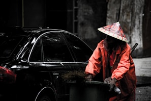 A person wearing an orange uniform and a conical hat is pushing a green trash bin filled with debris. The background shows a dark-colored car and a dim urban environment, possibly a street or alley.