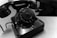 A black vintage rotary telephone with a circular dial displaying numbers from 0 to 9, resting on a wooden table. The phone's glossy surface reflects light, and its cord is visible on the side.