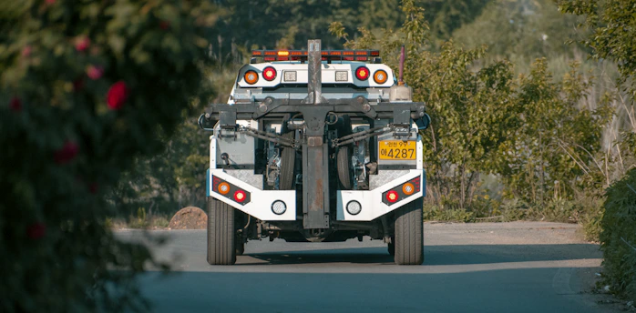 A tow truck is seen from behind on a paved road, surrounded by lush greenery. The truck has distinct red and orange lights and a license plate with numbers visible. The foreground features some blurred foliage.