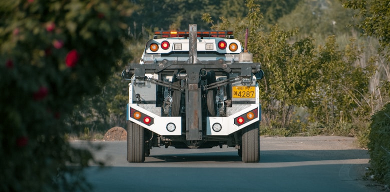 A tow truck is seen from behind on a paved road, surrounded by lush greenery. The truck has distinct red and orange lights and a license plate with numbers visible. The foreground features some blurred foliage.