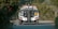A tow truck is seen from behind on a paved road, surrounded by lush greenery. The truck has distinct red and orange lights and a license plate with numbers visible. The foreground features some blurred foliage.