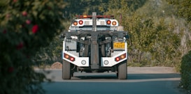 A tow truck is seen from behind on a paved road, surrounded by lush greenery. The truck has distinct red and orange lights and a license plate with numbers visible. The foreground features some blurred foliage.