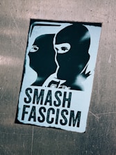 A sticker featuring two silhouetted figures wearing masks, depicted in a stencil art style. Bold text underneath reads 'Smash Fascism', indicating an anti-fascist message.