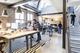 A modern office space with several people working on computers at shared desks. The room features exposed brick walls, large windows allowing natural light, and industrial-style lighting fixtures. A staircase on the right leads to an upper level while a man is descending. The decor includes striped black and white rugs under the desks.