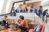 A modern office space with multiple people engaged in various activities. In the foreground, a man and a woman are having an animated conversation on a brown leather sofa. A laptop and a bottle are on the table in front of them. The background features a group of people standing and interacting near a brick wall, with one person using a laptop and others appearing to be in discussion. The setting is bright and open, with a contemporary industrial design featuring exposed brick and large windows. There are hanging ceiling lamps and a small table with pastries placed on it.