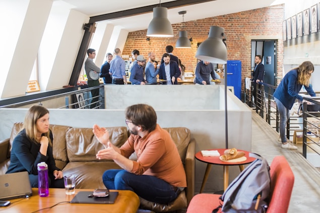 A modern office space with multiple people engaged in various activities. In the foreground, a man and a woman are having an animated conversation on a brown leather sofa. A laptop and a bottle are on the table in front of them. The background features a group of people standing and interacting near a brick wall, with one person using a laptop and others appearing to be in discussion. The setting is bright and open, with a contemporary industrial design featuring exposed brick and large windows. There are hanging ceiling lamps and a small table with pastries placed on it.