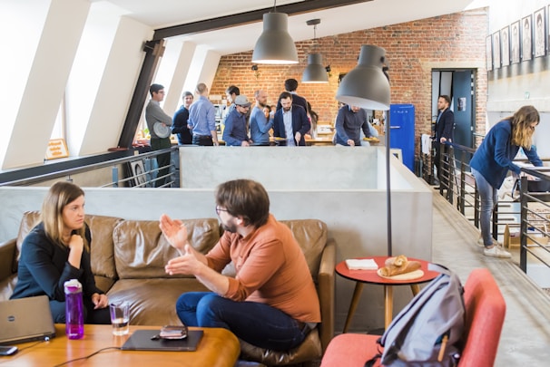 A modern office space with multiple people engaged in various activities. In the foreground, a man and a woman are having an animated conversation on a brown leather sofa. A laptop and a bottle are on the table in front of them. The background features a group of people standing and interacting near a brick wall, with one person using a laptop and others appearing to be in discussion. The setting is bright and open, with a contemporary industrial design featuring exposed brick and large windows. There are hanging ceiling lamps and a small table with pastries placed on it.