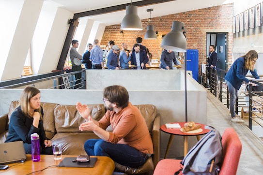 A modern office space with multiple people engaged in various activities. In the foreground, a man and a woman are having an animated conversation on a brown leather sofa. A laptop and a bottle are on the table in front of them. The background features a group of people standing and interacting near a brick wall, with one person using a laptop and others appearing to be in discussion. The setting is bright and open, with a contemporary industrial design featuring exposed brick and large windows. There are hanging ceiling lamps and a small table with pastries placed on it.