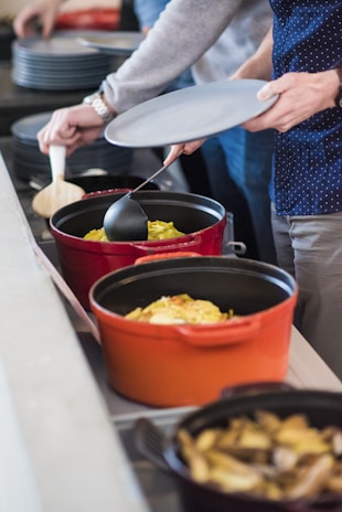 Several people are serving themselves food from a buffet with large pots. One person holds a gray plate while using a ladle to take food from an orange pot. Other pots and serving utensils are visible, with food such as noodles or pasta being served.