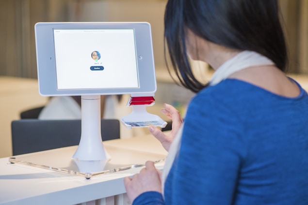 A woman interacts with a digital check-in kiosk that features a tablet screen displaying a user's profile image and a 'Confirm' button. She seems to be verifying her identity or checking in for an event or appointment. The setting suggests a modern office or reception area.