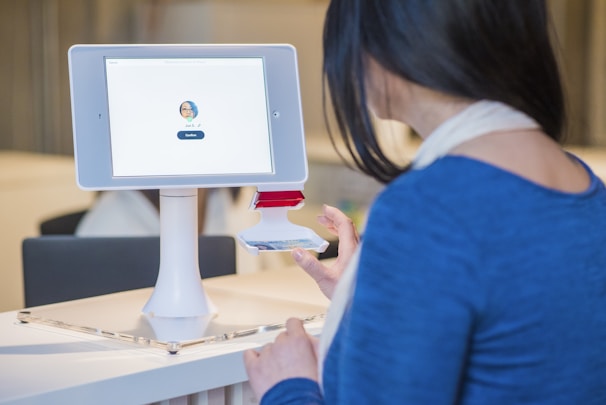 A woman interacts with a digital check-in kiosk that features a tablet screen displaying a user's profile image and a 'Confirm' button. She seems to be verifying her identity or checking in for an event or appointment. The setting suggests a modern office or reception area.