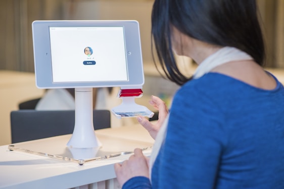 A woman interacts with a digital check-in kiosk that features a tablet screen displaying a user's profile image and a 'Confirm' button. She seems to be verifying her identity or checking in for an event or appointment. The setting suggests a modern office or reception area.