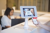 A person is holding a smartphone displaying a QR code towards a tablet mounted on a stand. The tablet screen shows a scan of the QR code. In the background, there is a blurred figure seated at a desk working on a laptop.