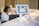 A person is holding a smartphone displaying a QR code towards a tablet mounted on a stand. The tablet screen shows a scan of the QR code. In the background, there is a blurred figure seated at a desk working on a laptop.