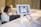 A person is holding a smartphone displaying a QR code towards a tablet mounted on a stand. The tablet screen shows a scan of the QR code. In the background, there is a blurred figure seated at a desk working on a laptop.