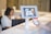 A person is holding a smartphone displaying a QR code towards a tablet mounted on a stand. The tablet screen shows a scan of the QR code. In the background, there is a blurred figure seated at a desk working on a laptop.