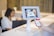 A person is holding a smartphone displaying a QR code towards a tablet mounted on a stand. The tablet screen shows a scan of the QR code. In the background, there is a blurred figure seated at a desk working on a laptop.