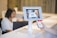 A person is holding a smartphone displaying a QR code towards a tablet mounted on a stand. The tablet screen shows a scan of the QR code. In the background, there is a blurred figure seated at a desk working on a laptop.