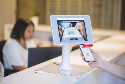 A person is holding a smartphone displaying a QR code towards a tablet mounted on a stand. The tablet screen shows a scan of the QR code. In the background, there is a blurred figure seated at a desk working on a laptop.