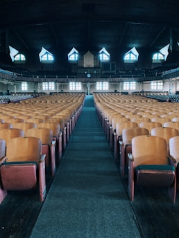 A spacious auditorium with rows of wooden seats leading to a stage. The ceiling is high and dark, with distinctively shaped windows allowing light to enter. The stage features a large organ, and the ambiance feels empty and historic.