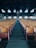 A spacious auditorium with rows of wooden seats leading to a stage. The ceiling is high and dark, with distinctively shaped windows allowing light to enter. The stage features a large organ, and the ambiance feels empty and historic.