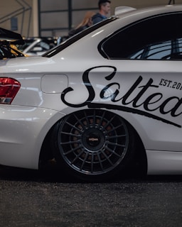 A sleek white car with a prominent black decal reading 'Salted' and 'EST. 2020' on the side. The car features dark multi-spoke wheels and is parked indoors, possibly at an auto show or event. There are blurred figures in the background, indicating other people are present.