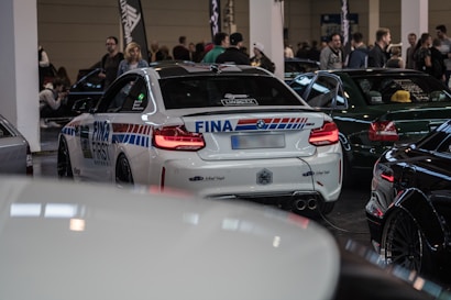 A car exhibition with several high-performance vehicles, prominently featuring a white sports car with racing decals and branding. Numerous people are seen in the background, suggesting a busy and vibrant event atmosphere. The setting appears to be indoors with artificial lighting.