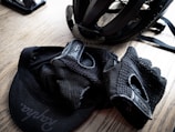 A cycling helmet and gloves are placed on a wooden surface, along with a cap featuring a visible logo. The items appear neatly arranged, suggesting preparedness for a cycling activity.