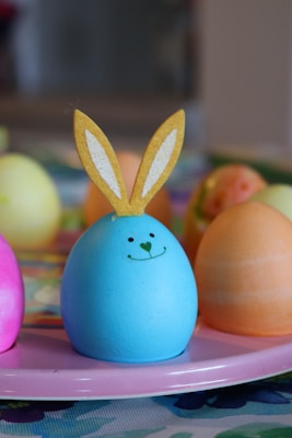 A pastel blue egg with a happy face drawn on it, topped with yellow felt bunny ears is centered on a pink plate. Surrounding the blue egg are other colorful eggs, including pink, yellow, and orange, on a patterned cloth with blurred colors in the background.