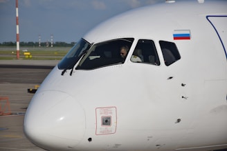 The close-up view of an airplane cockpit with the pilot visible inside. The fuselage is predominantly white, and a flag is displayed near the cockpit window. The airplane is positioned on an airport tarmac with runway elements visible in the background.