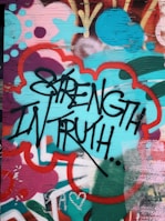 Graffiti art featuring bold black lettering of the words 'Strength in Truth' against a vibrant, multicolored background. The background includes abstract shapes and patterns in shades of teal, red, pink, and orange, creating a dynamic and energetic visual effect. The surface appears to be a wooden panel with visible grain.
